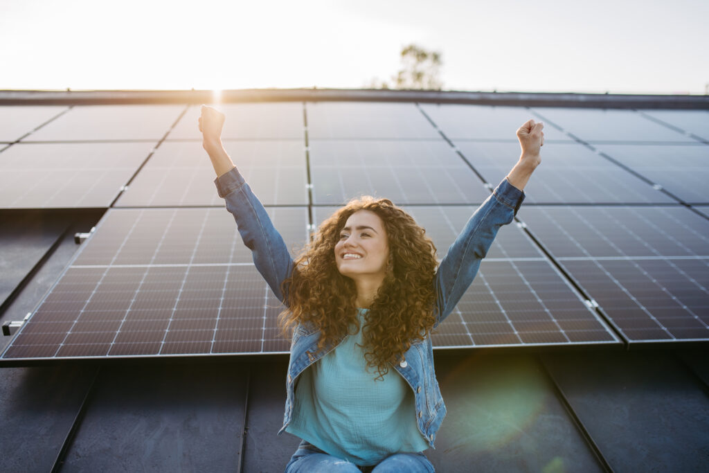 Weiblich gelesene Person streckt freudig Hände in die Höhe vor Dach mit Solarpanelen